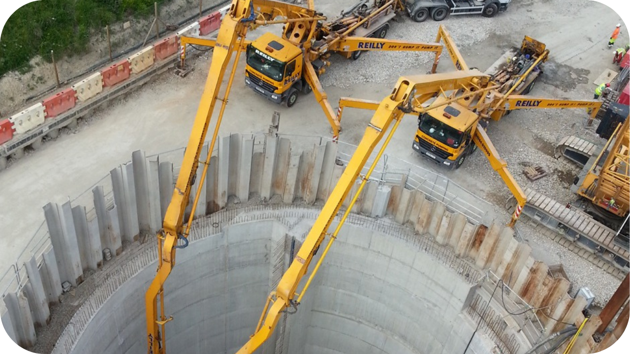 An aerial view of two large, yellow concrete boom pumps with their arms extending down into a deep, circular foundation.