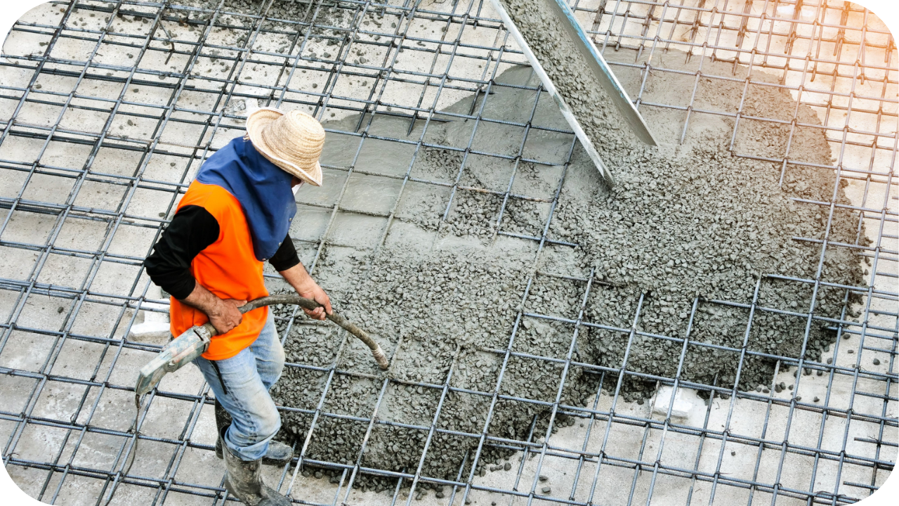 A worker in a straw hat uses a concrete vibrator to settle wet concrete being poured onto a large rebar grid.