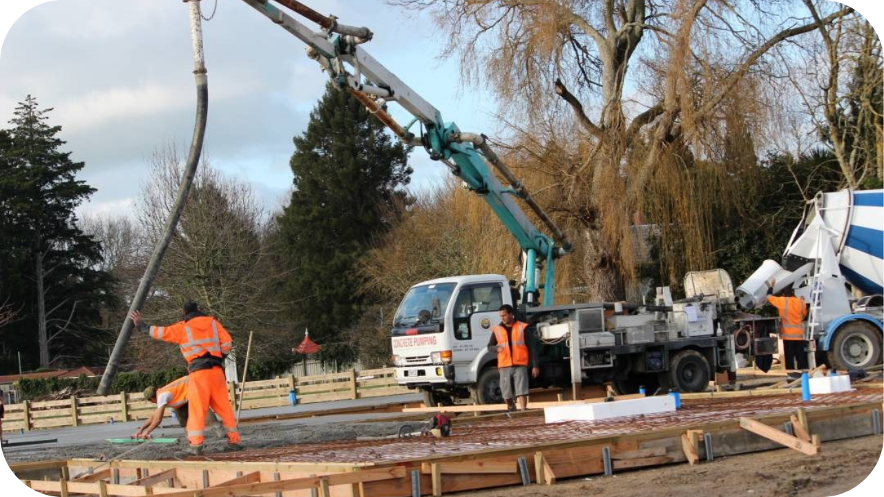 A concrete boom pump truck with its arm extended, pouring a large slab with the help of several workers in orange safety gear.