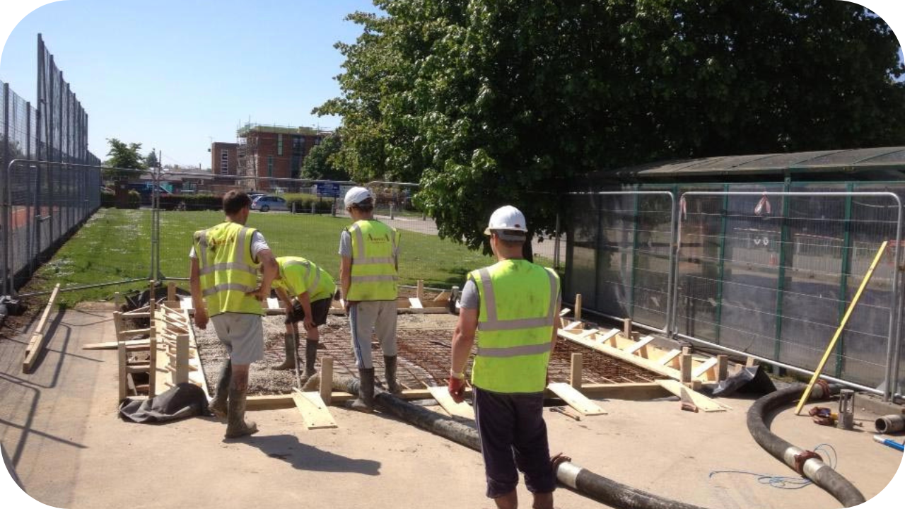 Construction crew in safety vests and boots managing a line pump hose while preparing foundations at an outdoor building site.