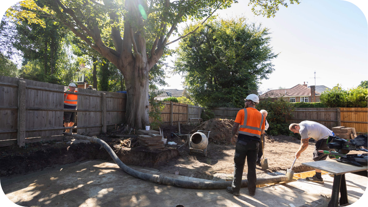 Construction workers operate a line pump in a backyard site, pouring concrete near a large tree with residential homes in the background.