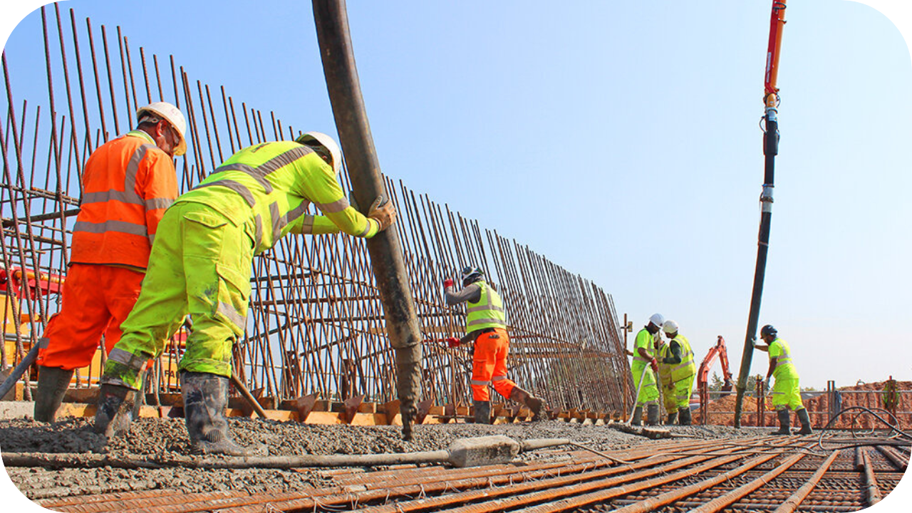 Construction workers in safety gear are pouring concrete with a pump hose beside steel reinforcement bars on site.