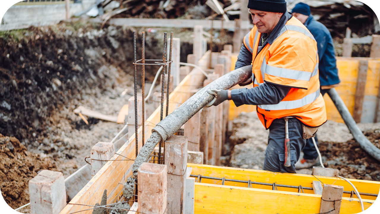 Worker in orange safety gear guiding concrete through a hose into timber formwork on a construction site foundation.