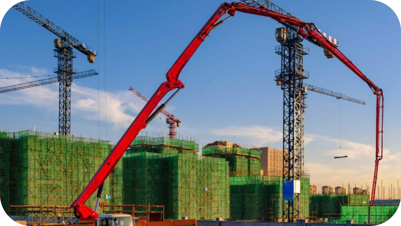 Large red boom pump extending across a construction site with cranes and green netted buildings during sunset.
