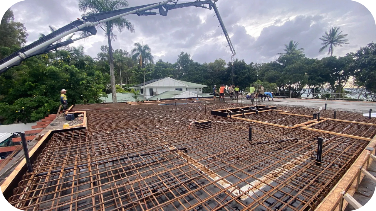 Workers pouring concrete at a construction site with a boom pump reaching over the steel reinforcement on a large foundation slab.