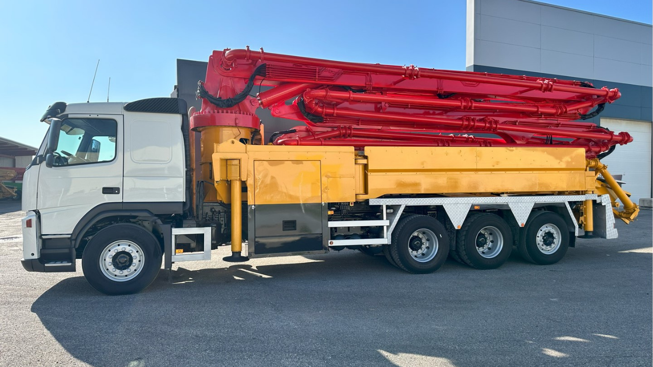 Side view of a white Volvo truck carrying a folded red concrete pump boom and yellow pump unit parked at a Melbourne work yard.