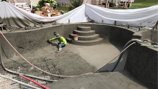 Worker smoothing fresh concrete inside a curved residential pool shell as pumping hoses deliver mix across the site during construction.