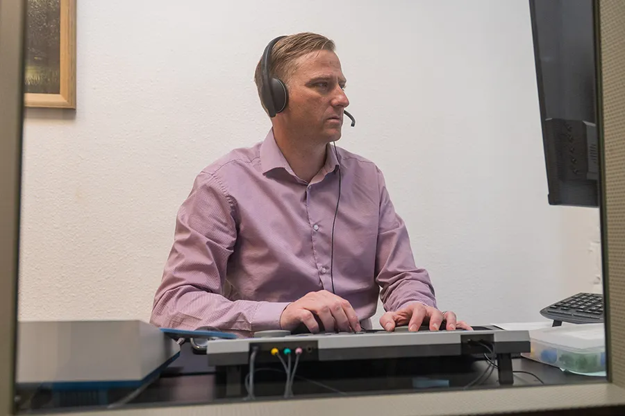 Male audiologist in a purple shirt wearing a headset, typing on a keyboard while looking at a computer monitor.