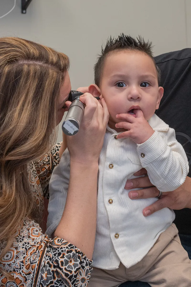 Female audiologist using an otoscope to examine a child's ear while the child sits calmly with fingers near his mouth.