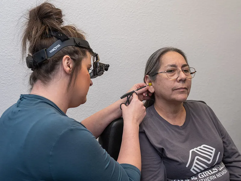 Female audiologist wearing magnifying headset examines the ear of a seated woman with glasses and a gray Boys & Girls Clubs shirt.
