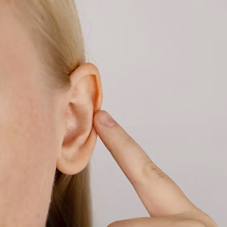 Close-up of a person touching the outer ear with their index finger against a plain background.