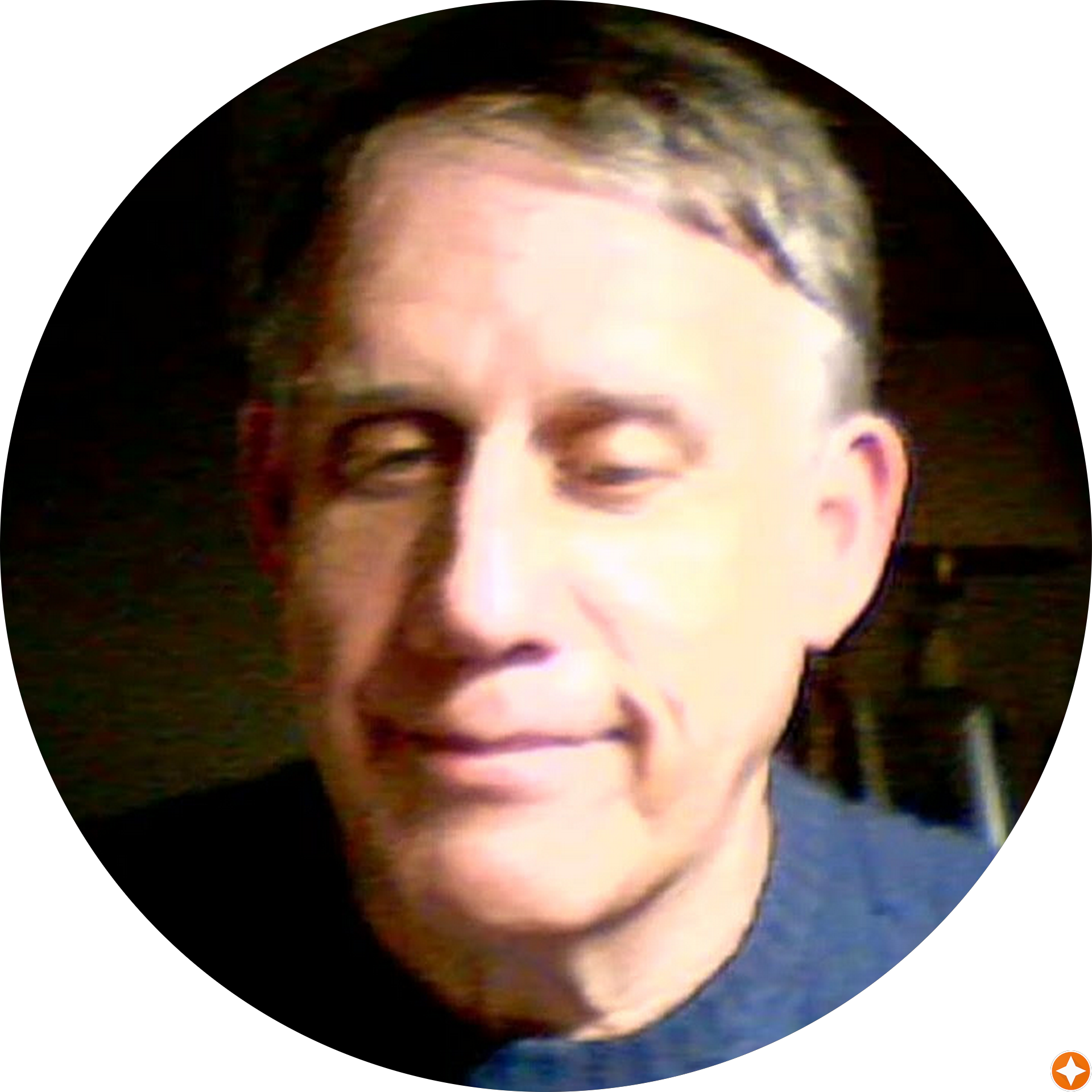 Bald man wearing a short-sleeve button-up shirt sitting outdoors on a light-colored bench with a wooden railing and trees in the background.