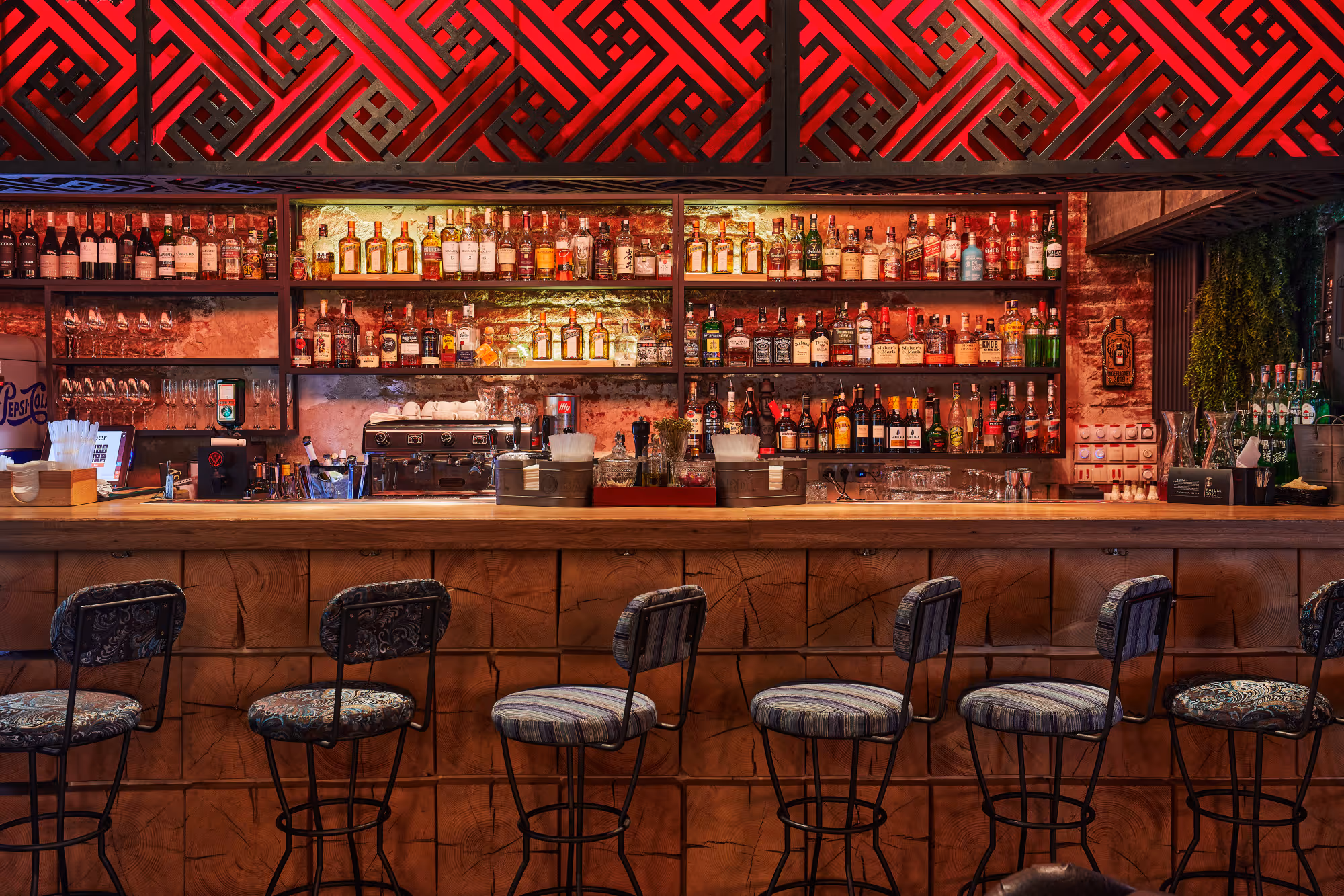 Bar with wooden counter, six stools, shelves stocked with liquor bottles under red patterned ceiling light.