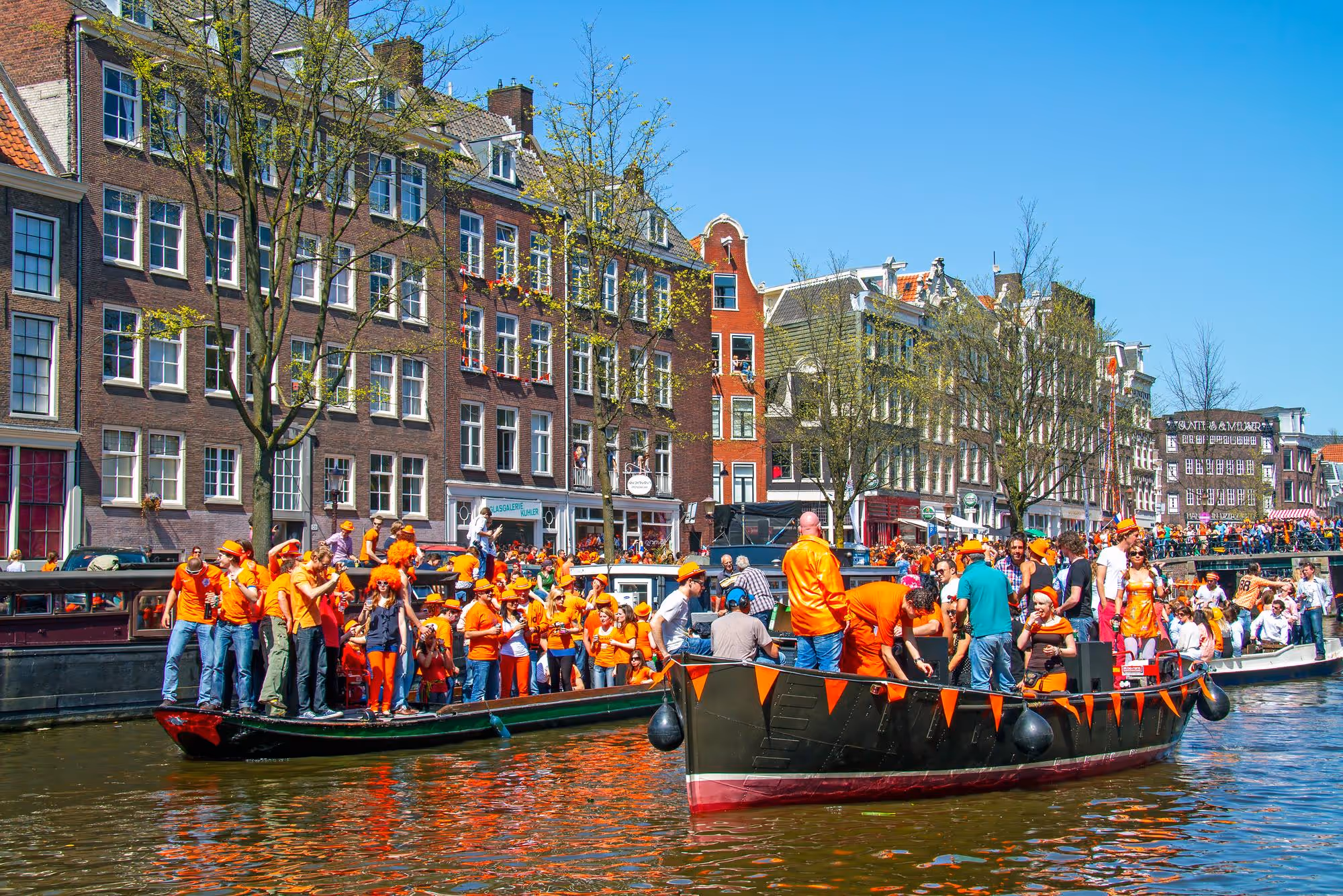 Crowd wearing orange on boats and canal in Amsterdam under blue sky by tall traditional buildings.