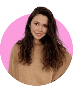 Smiling young woman with long curly hair wearing a light brown sweater in front of a pink circular background.