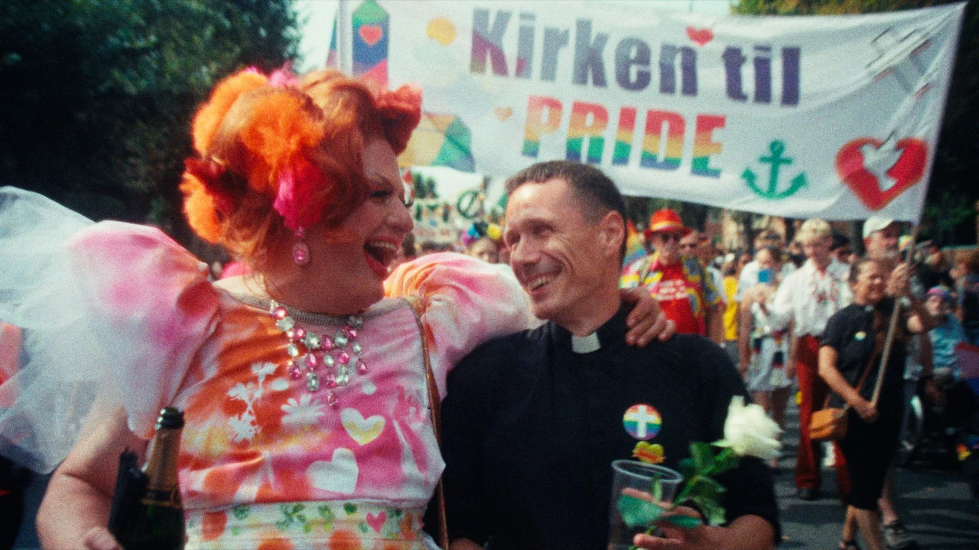 Pressebillede fra Alle Guds farver: En person i farverig drag og en præst foran et Kirken til Pride-banner.