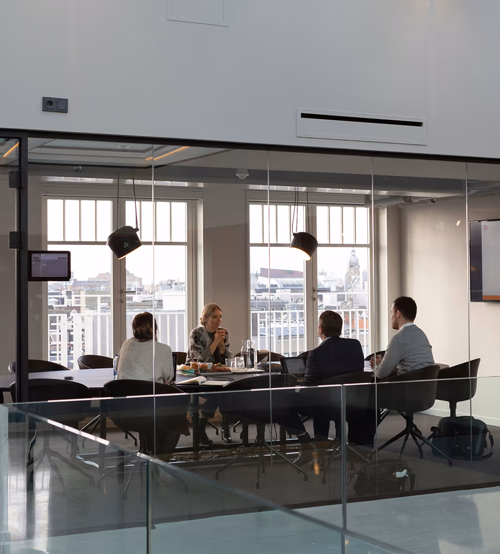 Four people seated around a conference table in a modern glass office meeting room with cityscape visible through large windows.