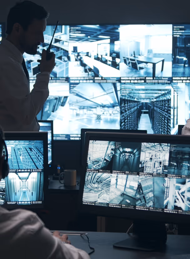 Security control room with multiple monitors displaying surveillance camera footage and a man speaking into a walkie-talkie.
