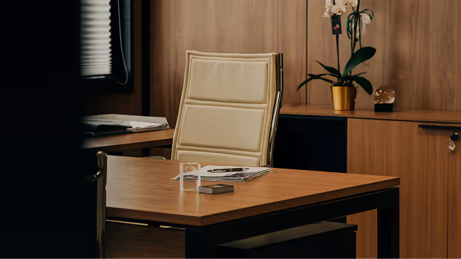 Modern office with a beige leather chair behind a wooden desk with papers and a pen, and a potted orchid on a cabinet in the background.