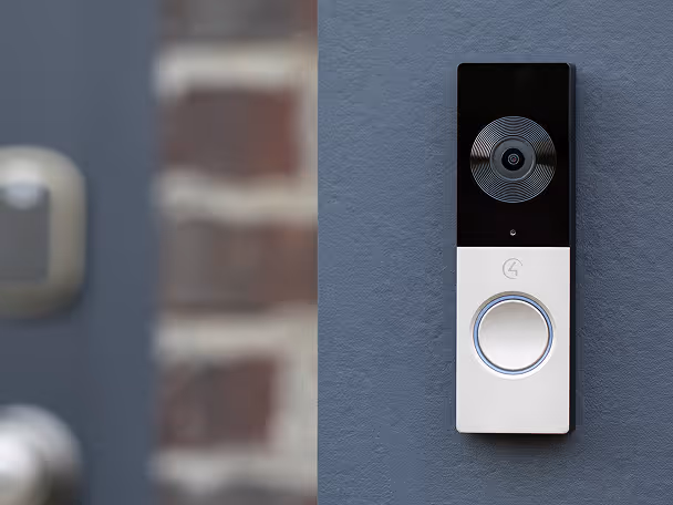 Modern video doorbell mounted on a gray wall beside a blurred door and brick wall background.