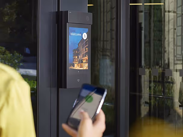 Person holding a smartphone near a modern access control system with a screen displaying 'Welcome' at a building entrance.