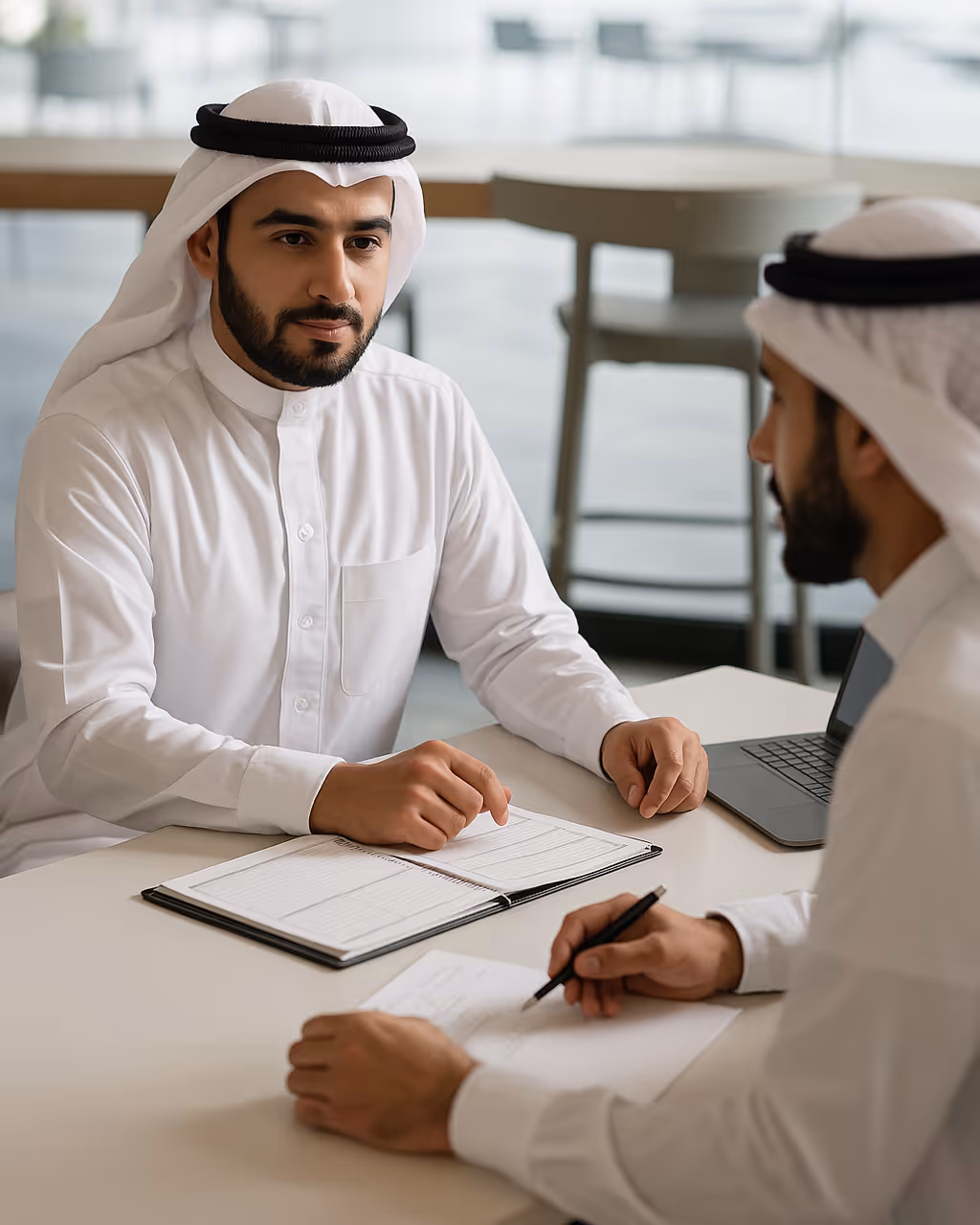 Two men in traditional Middle Eastern attire having a discussion at a table with a notebook, papers, and a laptop.
