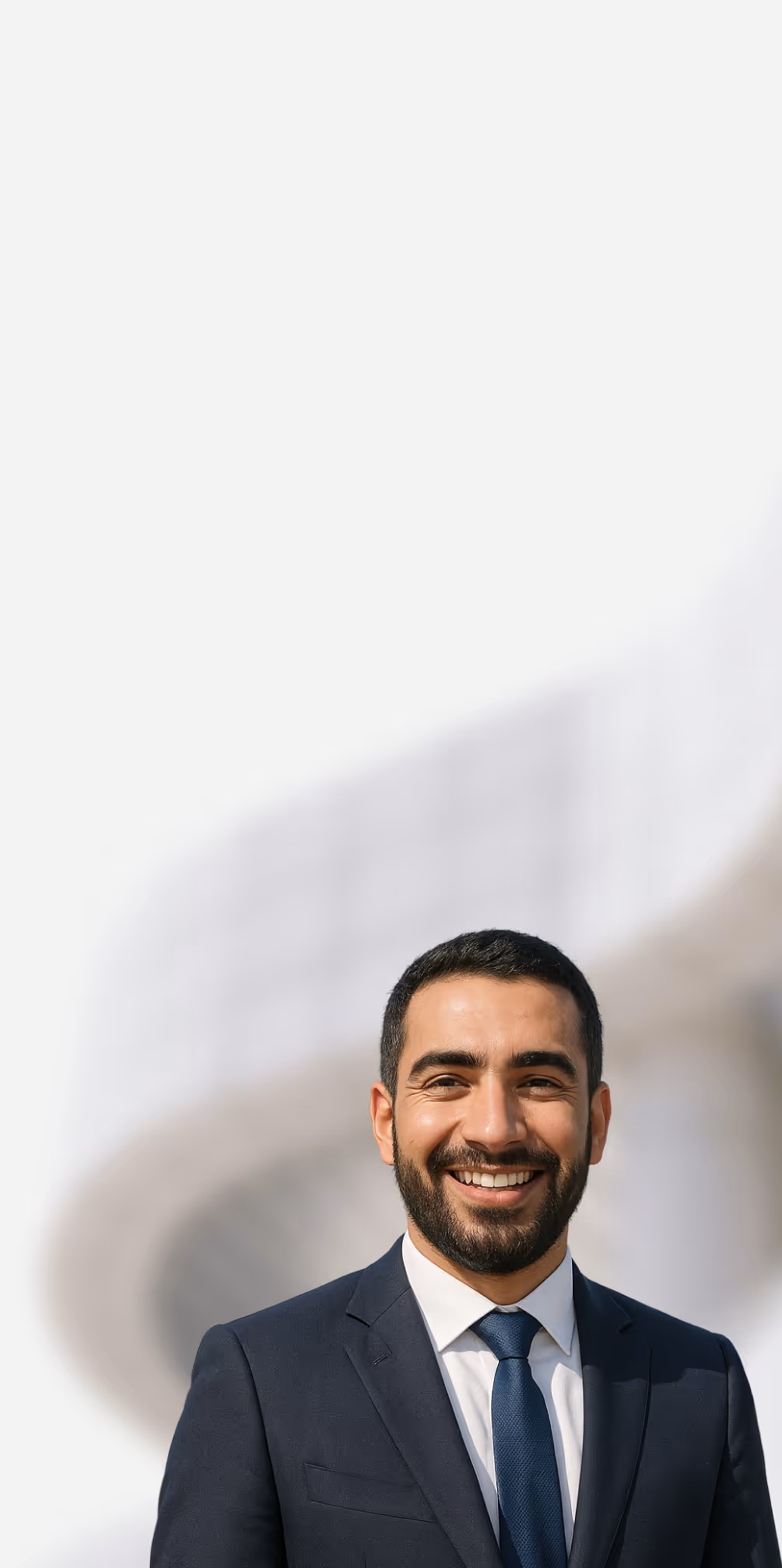 Smiling man with beard wearing a dark suit and blue tie standing outdoors with blurred modern architectural background.