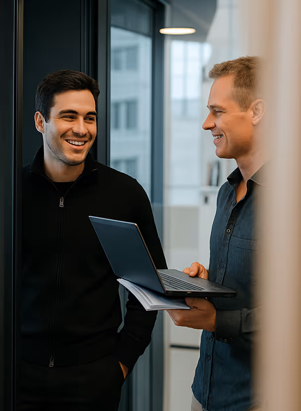 Two men standing and smiling while discussing something with one holding a laptop and papers in an office setting.