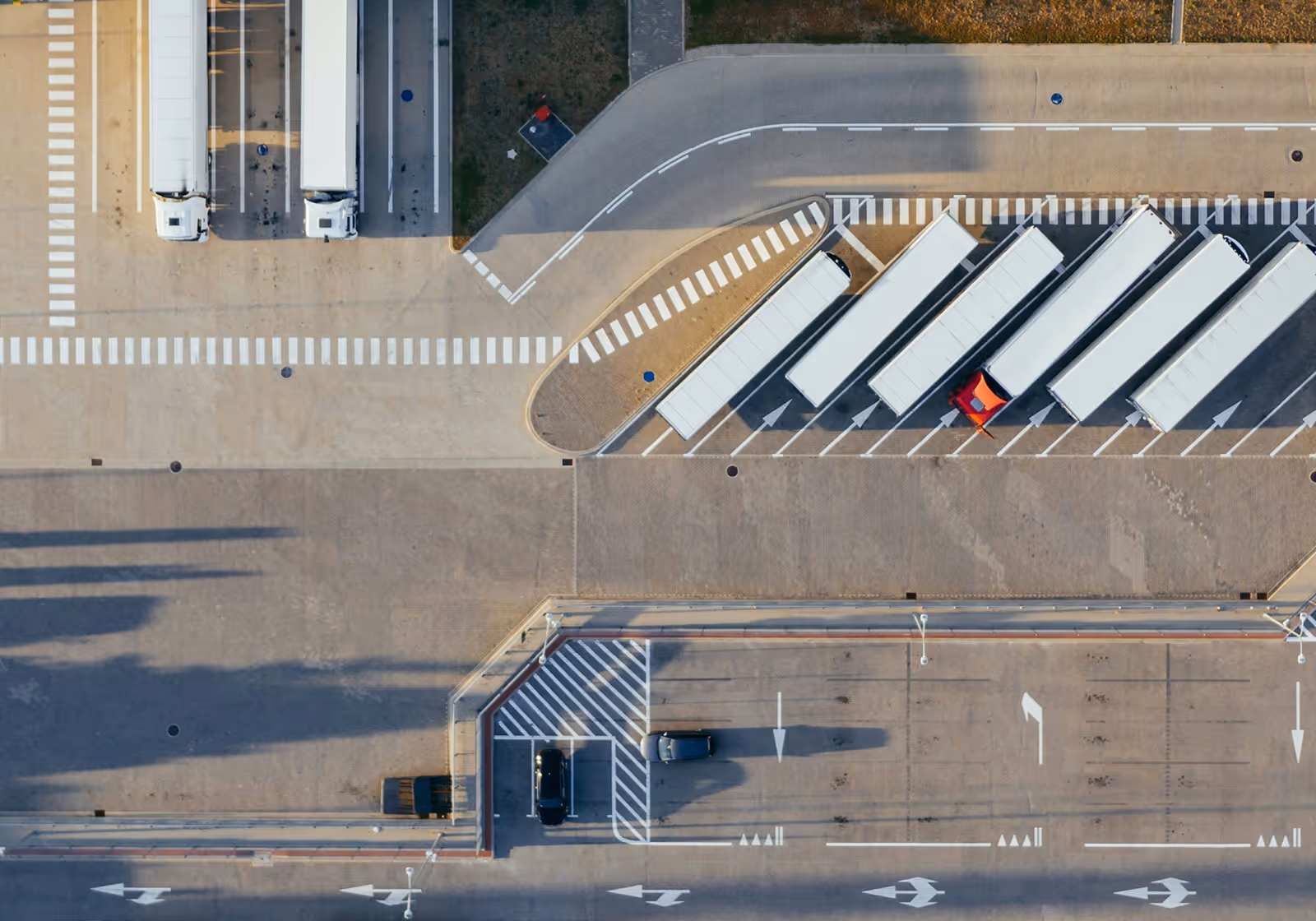 Aerial view of a parking area with several white trucks lined up and a few smaller vehicles parked and driving in marked lanes.