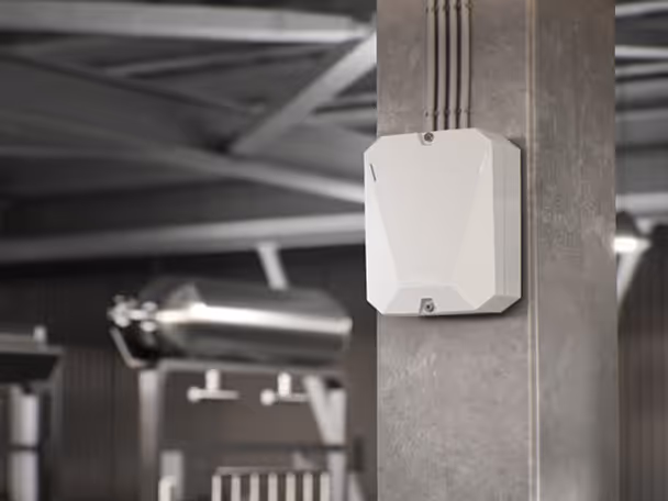 White rectangular industrial sensor or wireless device mounted on a concrete pillar with cables running above it in a factory setting.