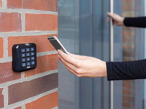 Hand holding a smartphone near a keypad access control on a brick wall beside a glass door.