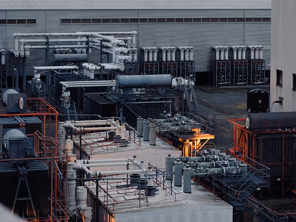 Industrial facility with interconnected pipes, tanks, and machinery against a metal building backdrop.