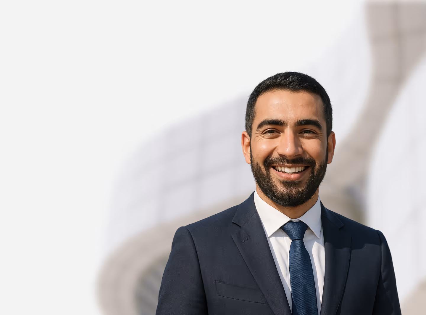 Smiling man with beard wearing a navy suit, white shirt, and blue tie standing outdoors.