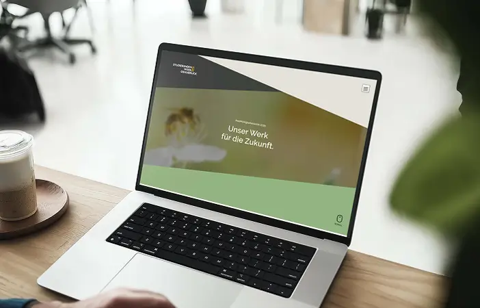 Laptop auf einem Holztisch mit geöffneter Webseite, die den Startscreen des Nachhaltigkeitsberichts mit dem Text 'Unser Werk für die Zukunft.' zeigt, daneben ein Kaffee und unscharfe Büromöbel im Hintergrund.