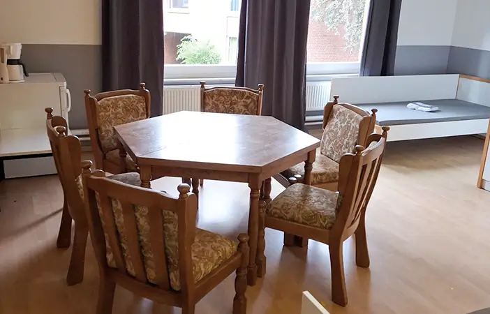 Sechseckiger Holztisch mit sechs gepolsterten Stühlen in einem hellen Raum mit Fenster, Vorhängen, Kühlschrank und einem Bett.