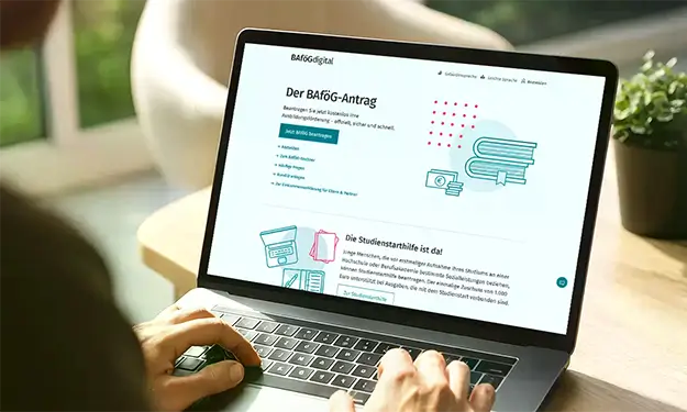 Laptop mit geöffneter Webseite zum BAföG-Antrag, auf dem Schreibtisch stehend, Hände auf Tastatur.