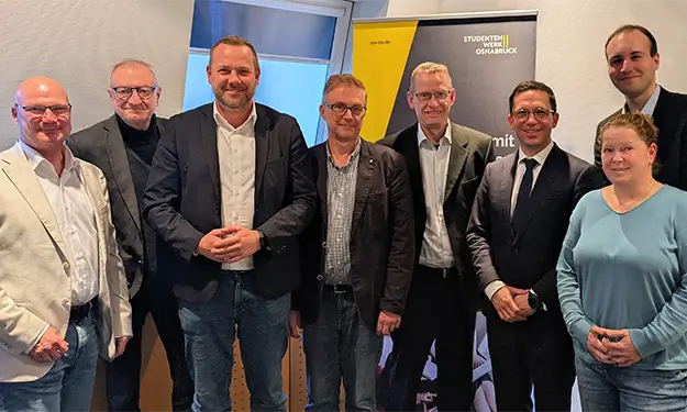 Gruppe von acht Personen steht in einem Besprechungsraum vor einem Banner mit Texten 'sw-os.de' und 'STUDIERENDENWERK OSNABRÜCK'.
