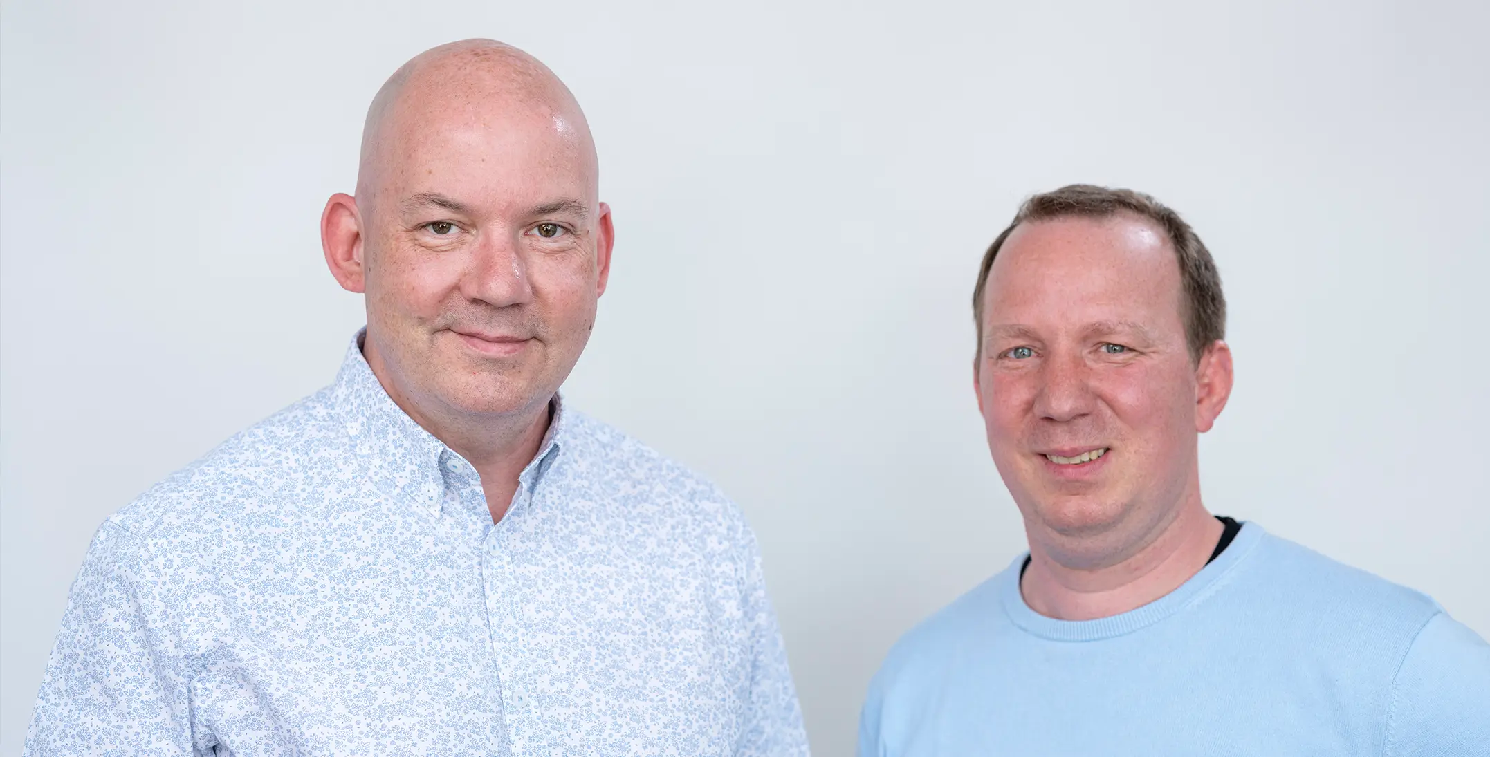 Marcus Stillwell und Martin Doll stehen vor einer hellen Wand und blicken lächelnd in die Kamera.