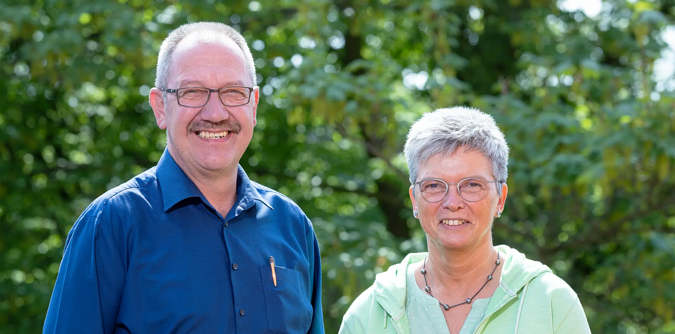 Rüdiger Hantke und Martina Marek stehen draußen nebeneinander vor einem grünen Busch und blicken in die Kamera.