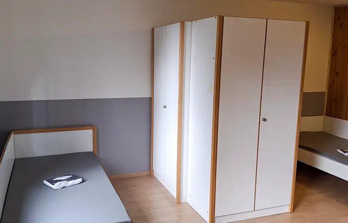 Ein einfach möbliertes Zimmer mit zwei Einzelbetten, die durch große weiße Schränke voneinander getrennt sind.