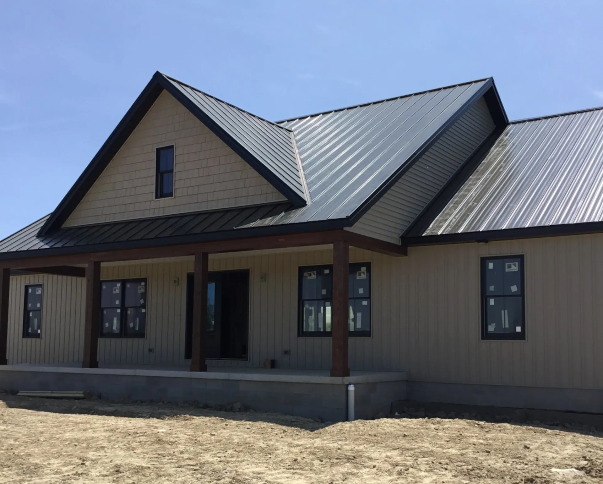 New home featuring a standing seam metal roof with black trim, board and batten siding, and covered front porch under construction on a clear sunny day.
