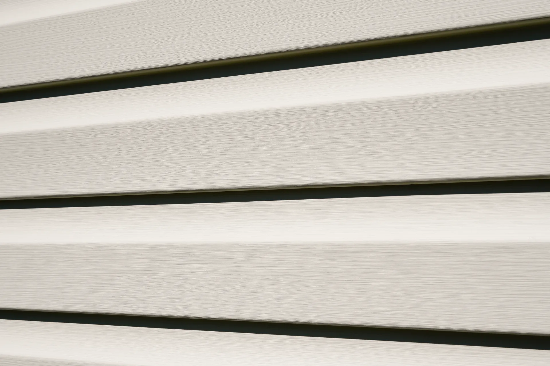 Close-up of beige vinyl siding panels arranged horizontally with narrow gaps between them, showing subtle texture and shadows.