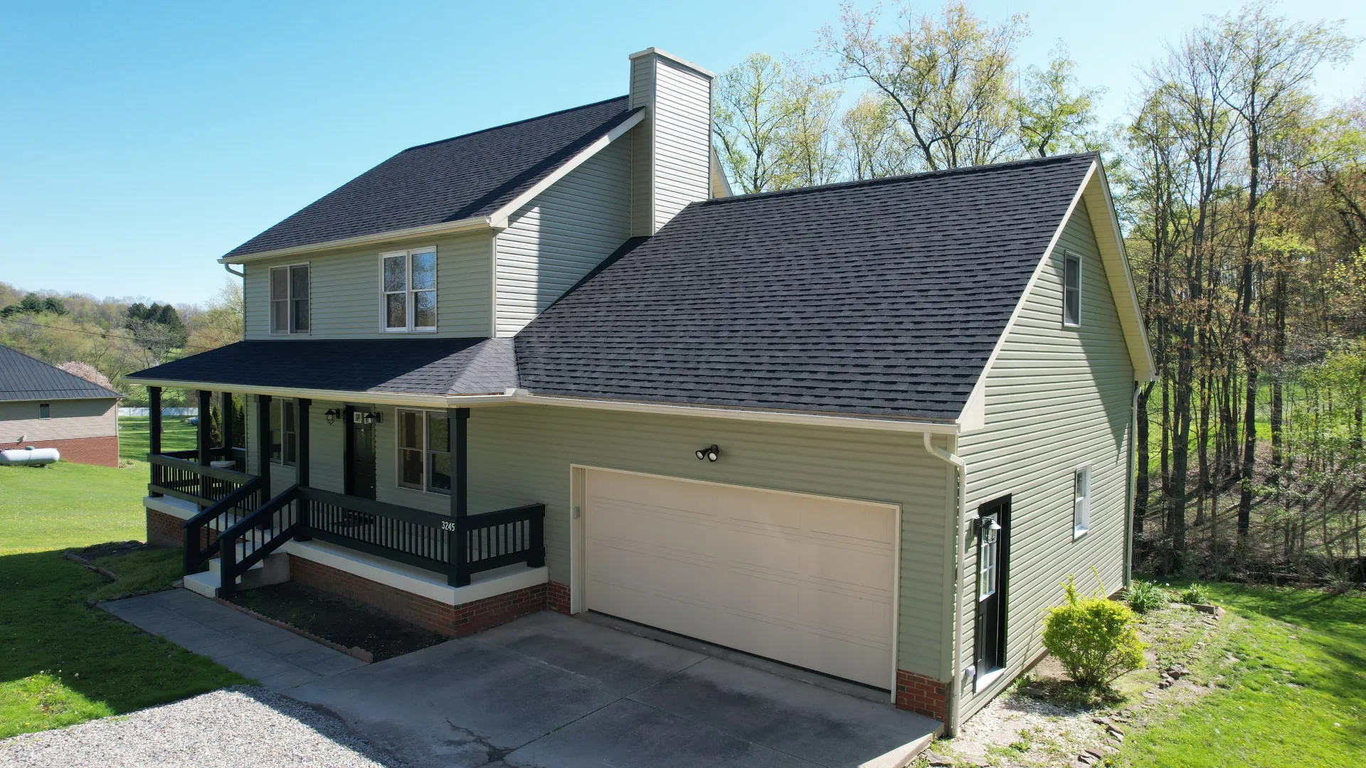 Two-story pale green house with a large attached garage and a black asphalt shingle roof, front porch with black railing, surrounded by green yard and trees.