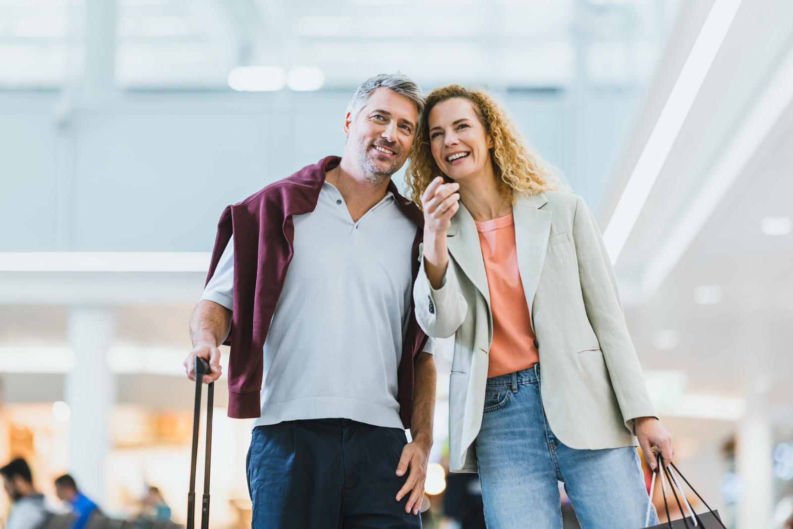 Markenfotografie: Lachendes Paar mit Rollkoffer beim Bummel durch die Shopping-Area am Flughafen München