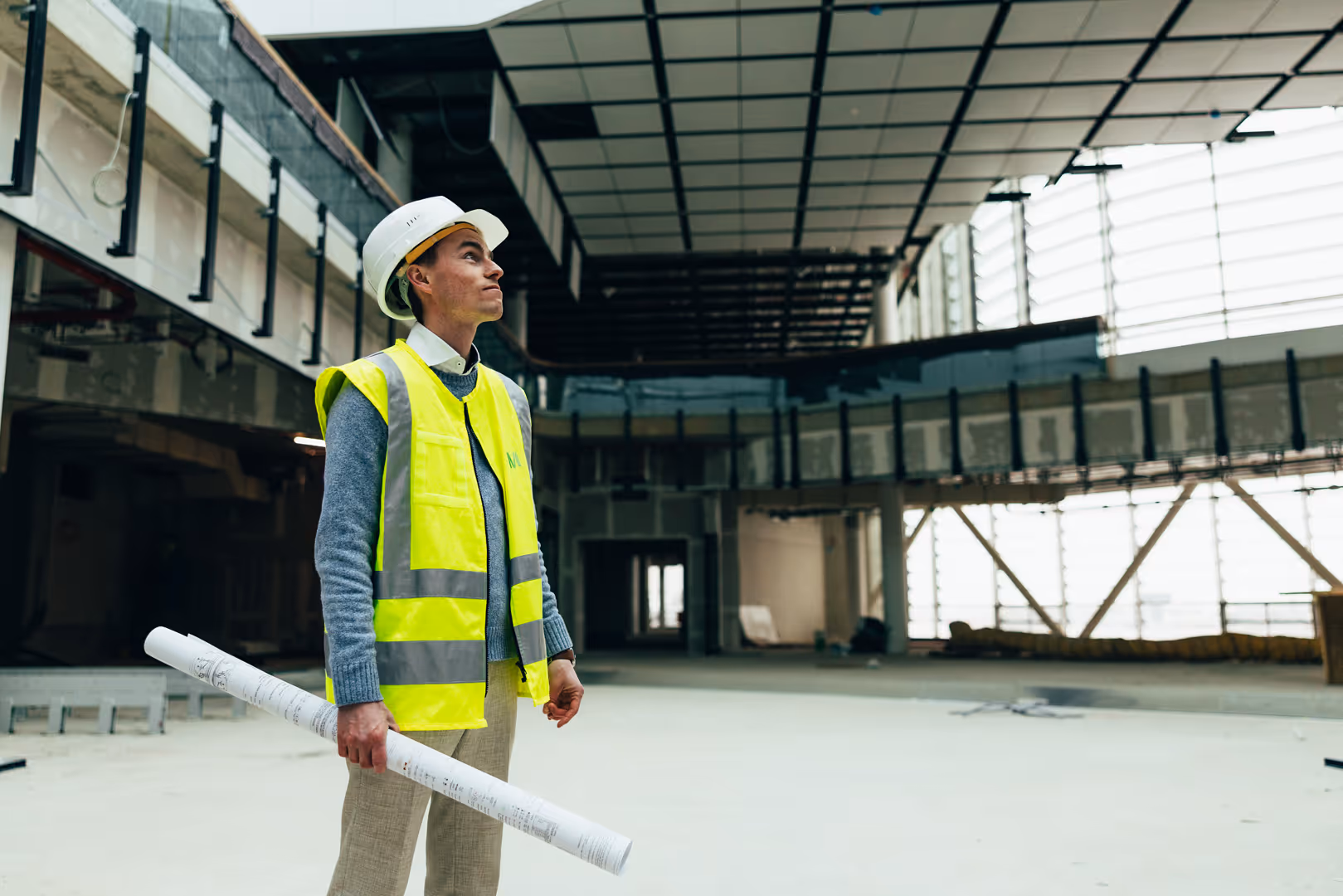 Mitarbeiterfotografie: Mann in Warnweste mit Bauplänen in einer Halle im Rohbau am Flughafen München