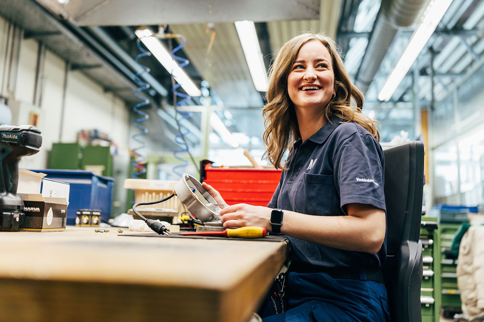 Mitarbeiterfotografie: Mitarbeiterin lächelt an der Werkbank in einer Werkstatt des Flughafens München