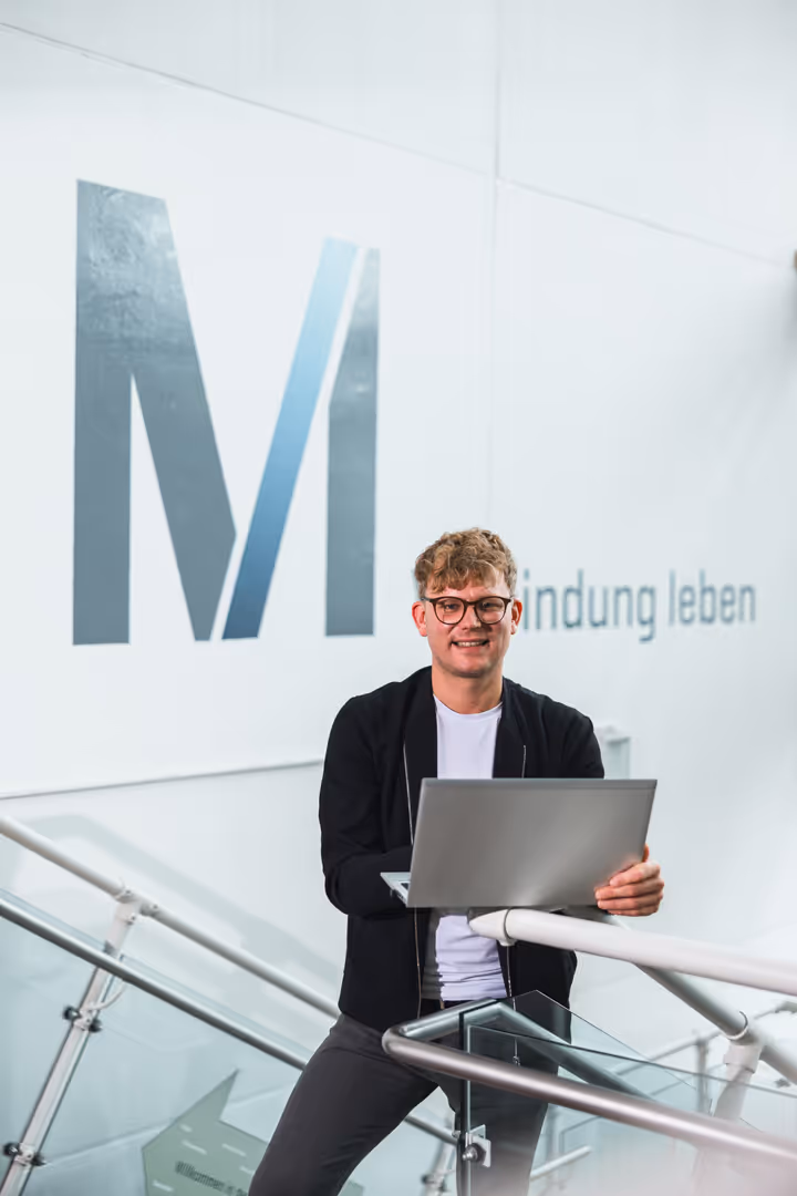 Mitarbeiterfotografie: Mitarbeiterin mit Laptop auf der Treppe vor dem großen M-Logo des Flughafens München