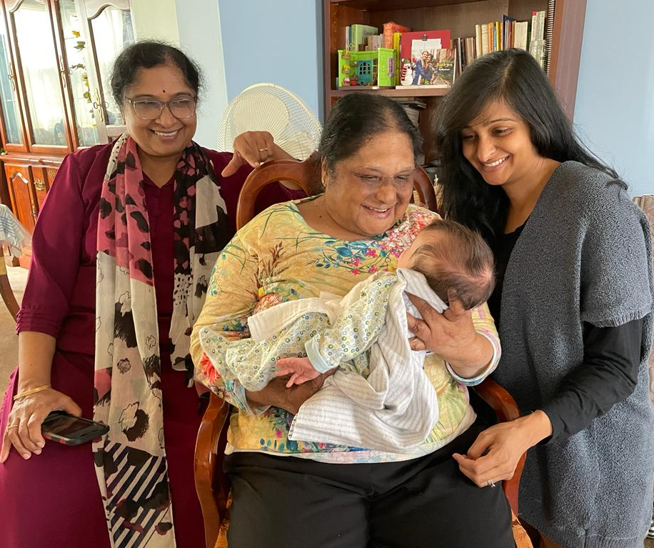 A picture of 3 adults and 1 child. The adult in the middle is holding a baby and everyone is looking at the baby and smiling.