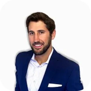 Smiling man with dark hair and beard wearing a white shirt and navy blazer against a plain white background.