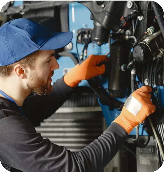 Mechanic in a blue cap and orange gloves working on a machine's engine components.